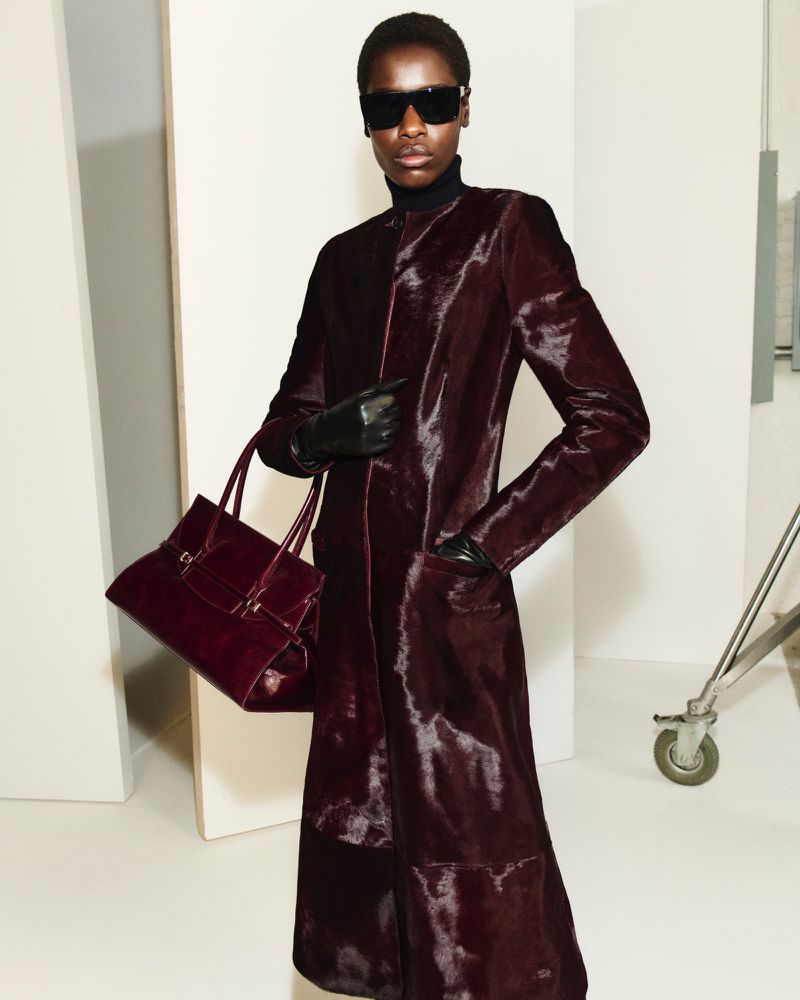 Elegant woman in a long maroon coat, sunglasses, and gloves carrying a matching handbag in a minimalist studio setting.