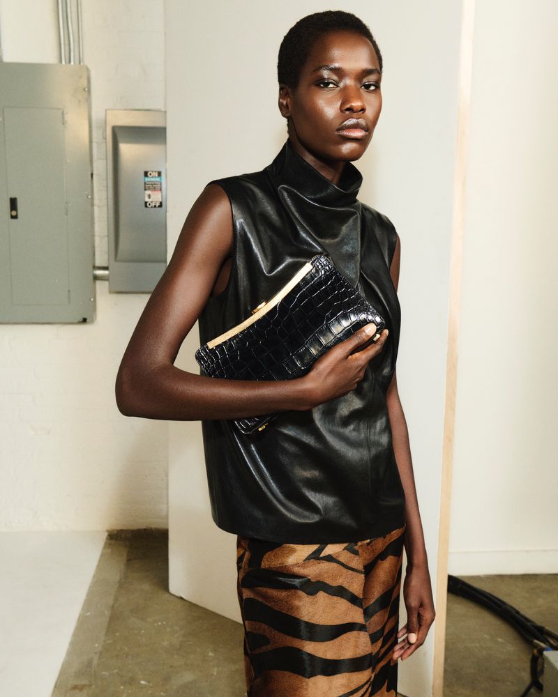 Elegant dark-skinned woman in a black leather top holds a crocodile-patterned clutch bag in a modern indoor setting with neutral walls.