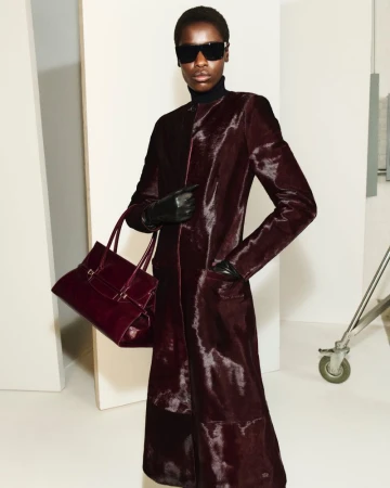 Model wearing a dark burgundy textured coat and sunglasses holding a matching handbag in a studio setting for Simkhai