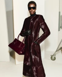 Model wearing a dark burgundy textured coat and sunglasses holding a matching handbag in a studio setting for Simkhai
