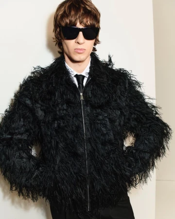 Model wearing a black feathered jacket and sunglasses against a light background for Simkhai