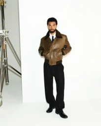 Man wearing a brown leather jacket and black trousers posing against a bright white studio backdrop with equipment.