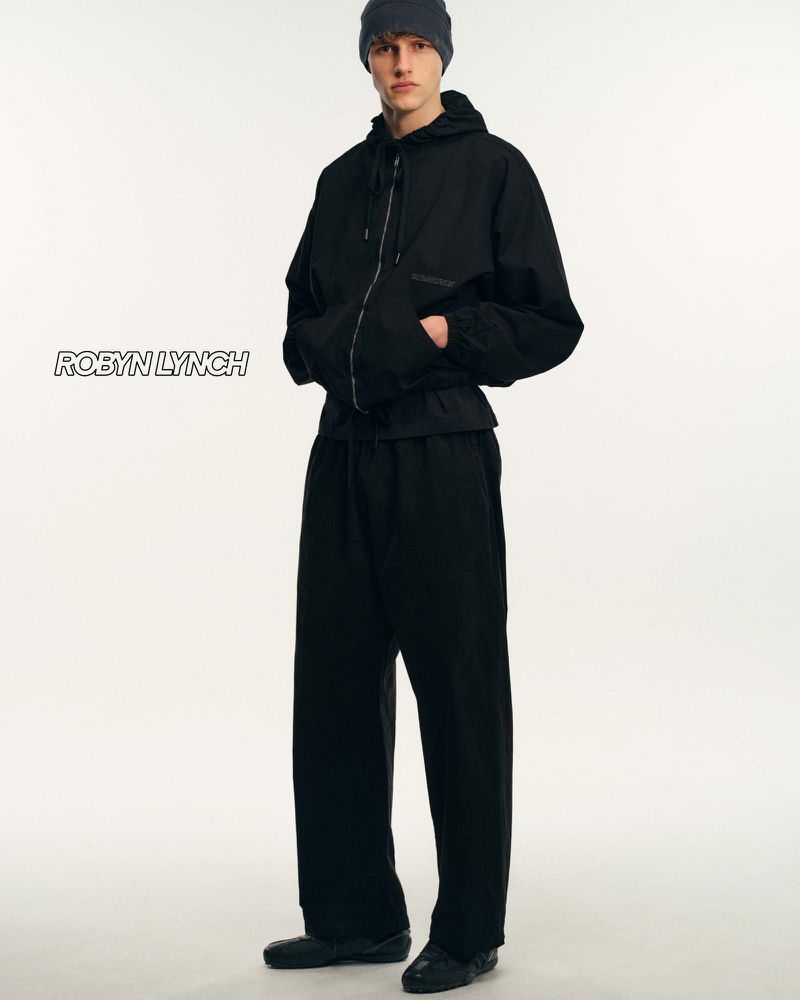 Male fashion model wears a black zip-up hooded jacket, matching wide-leg pants, and beanie against a white backdrop in a studio shot.