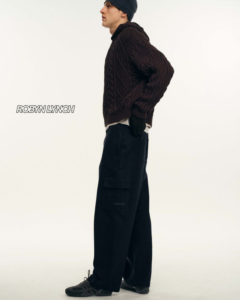 Fashionable adult male poses in a hooded knit sweater and wide-leg cargo pants against a white backdrop in a studio setting.