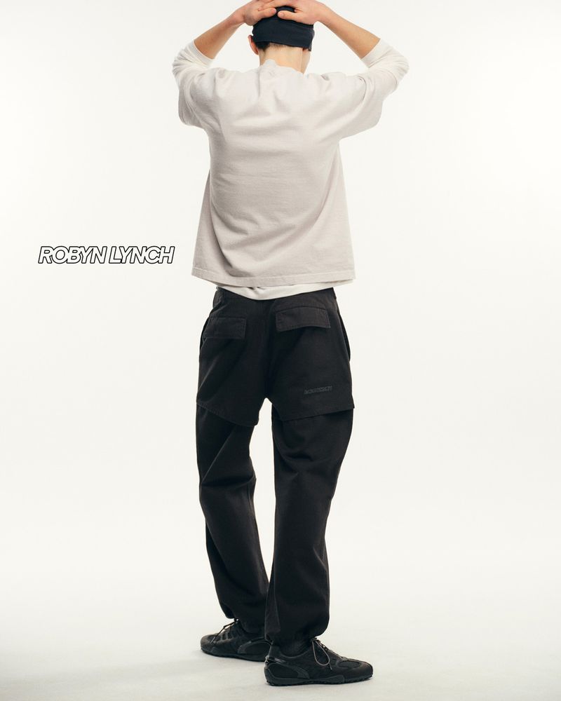 Rear view of a person in a light sweater, dark pants, and dark shoes posing against a white backdrop in a studio setting.