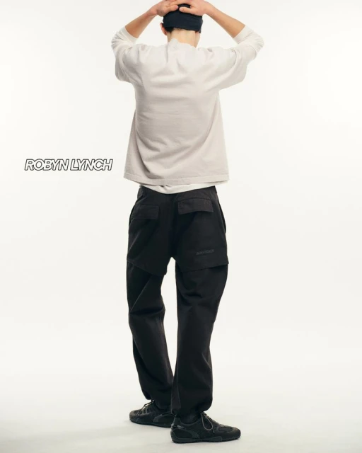 Rear view of a person in a light sweater, dark pants, and dark shoes posing against a white backdrop in a studio setting.