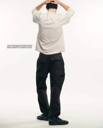 Rear view of a person in a light sweater, dark pants, and dark shoes posing against a white backdrop in a studio setting.