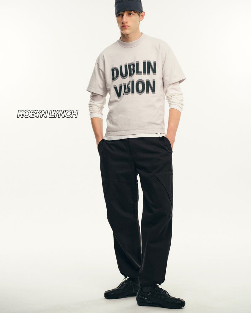 Fashion model wearing a grey tee shirt, black pants, and black shoes stands in a bright photo studio with a simple white background.