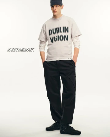 Fashion model wearing a grey tee shirt, black pants, and black shoes stands in a bright photo studio with a simple white background.