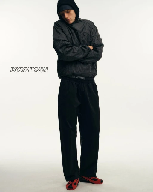 Full-length studio shot of a young adult man in a hooded jacket and sweatpants with arms crossed against a white background.