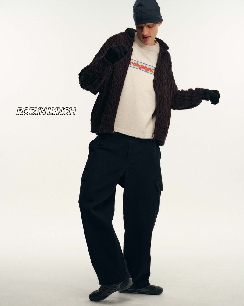 Full-length shot of an adult male model in a navy beanie, cable knit cardigan, t-shirt, and cargo pants, posing in a photo studio.