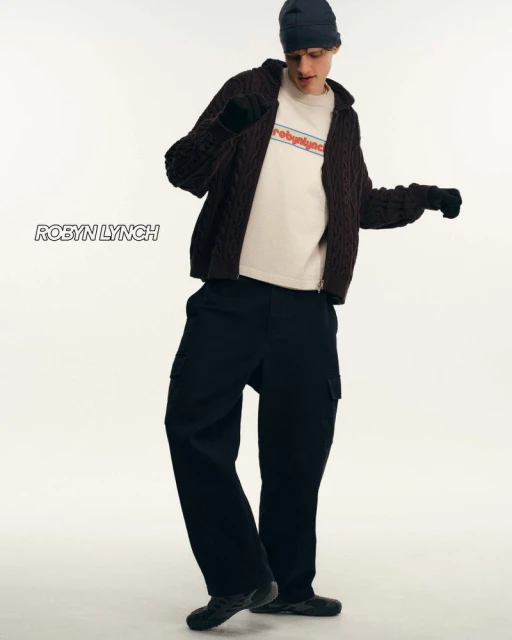 Full-length shot of an adult male model in a navy beanie, cable knit cardigan, t-shirt, and cargo pants, posing in a photo studio.