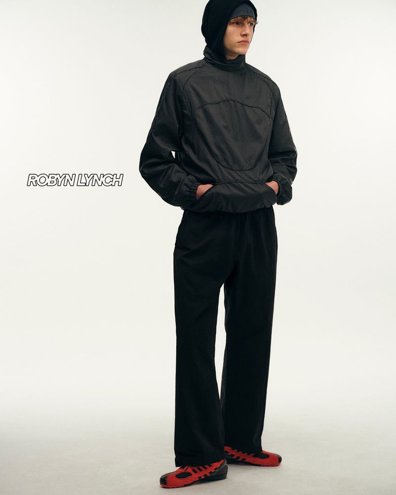 Full shot of male fashion model posing in black hooded jacket, wide-leg trousers, and red sneakers against a bright, neutral backdrop.