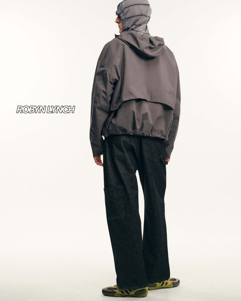 Model seen from behind wearing a grey hooded jacket and dark trousers against a plain white studio background.