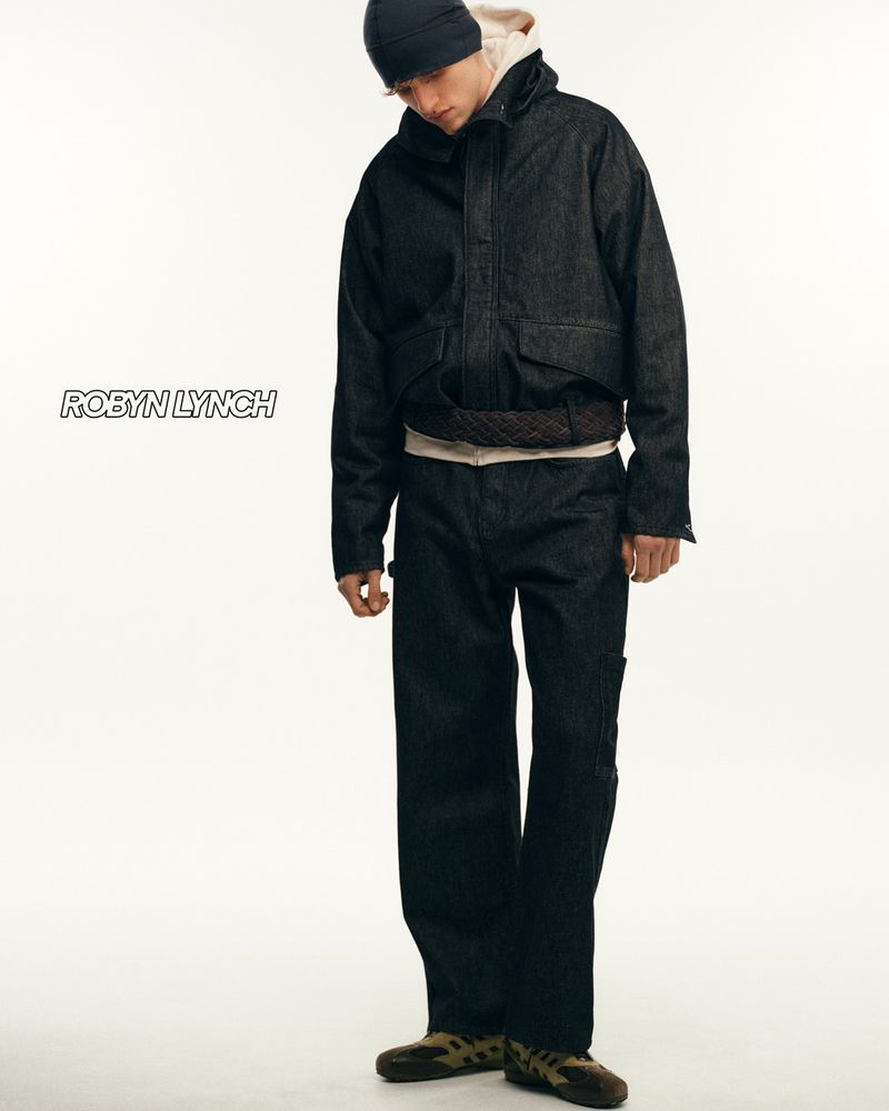 Fashion shot of a male model in a denim jacket, jeans and beanie, standing against a white background in a studio setting.