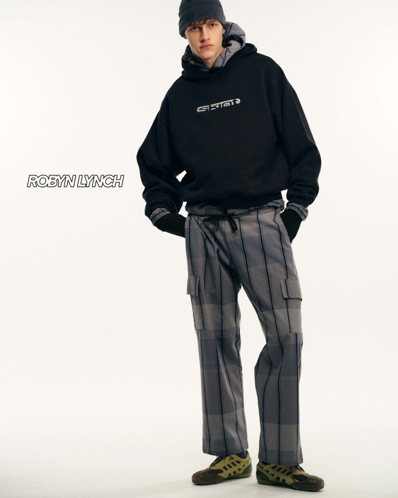 Adult male in a studio wearing a black hoodie and gray plaid pants, complemented by a gray beanie, against a clean white backdrop.