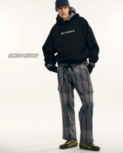 Adult male in a studio wearing a black hoodie and gray plaid pants, complemented by a gray beanie, against a clean white backdrop.