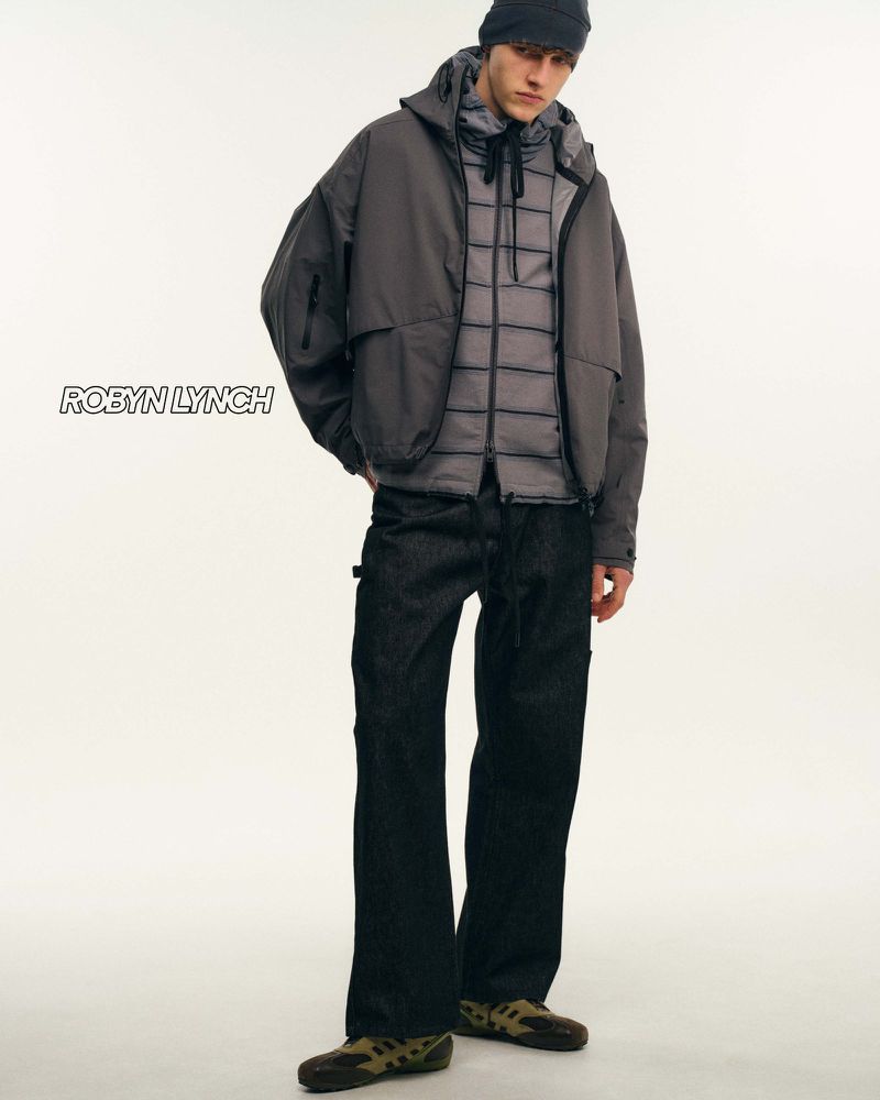 Male fashion model poses in hooded jacket, puffer vest, dark jeans, and moccasins against a stark white backdrop in a studio shot.