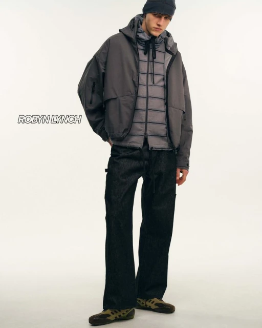 Male fashion model poses in hooded jacket, puffer vest, dark jeans, and moccasins against a stark white backdrop in a studio shot.