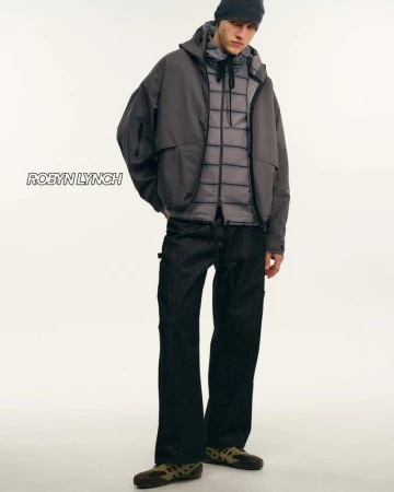 Male fashion model poses in hooded jacket, puffer vest, dark jeans, and moccasins against a stark white backdrop in a studio shot.
