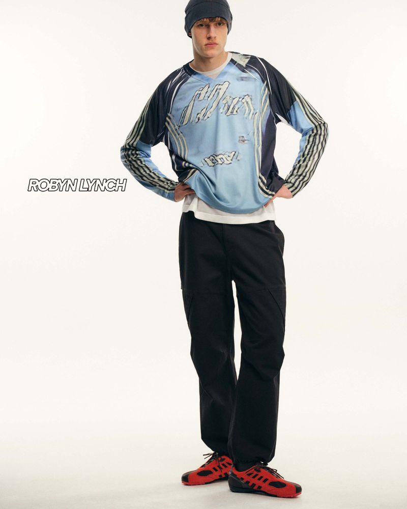 Full-length studio portrait of a young adult man wearing a long-sleeved graphic shirt, black pants, beanie, and red sneakers, on white.