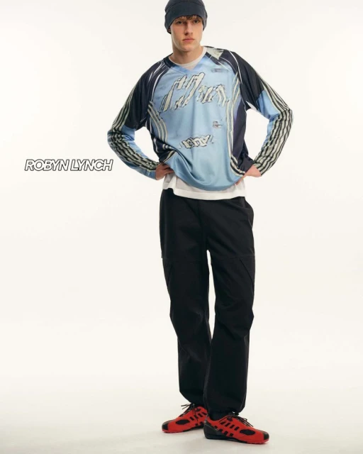 Full-length studio portrait of a young adult man wearing a long-sleeved graphic shirt, black pants, beanie, and red sneakers, on white.