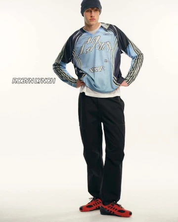 Full-length studio portrait of a young adult man wearing a long-sleeved graphic shirt, black pants, beanie, and red sneakers, on white.