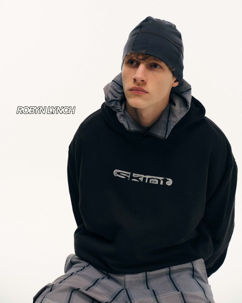 Young man in a black hoodie and gray patterned pants posing in a bright photo studio wearing a gray beanie-style hat.