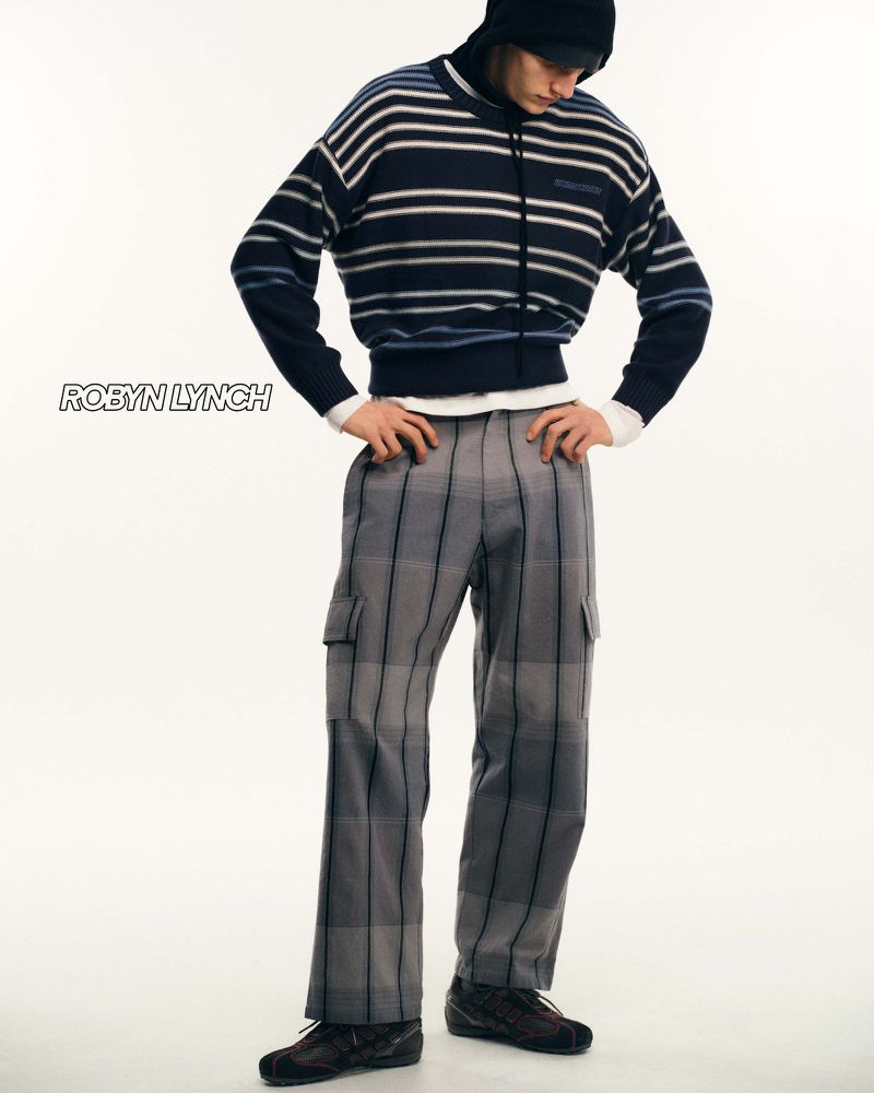 Male fashion model poses in a striped sweater and plaid pants in a full shot against a bright studio backdrop, showcasing contemporary menswear.