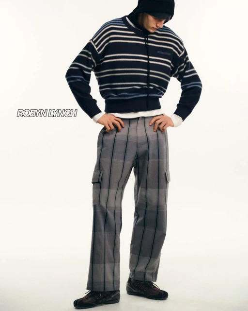 Male fashion model poses in a striped sweater and plaid pants in a full shot against a bright studio backdrop, showcasing contemporary menswear.