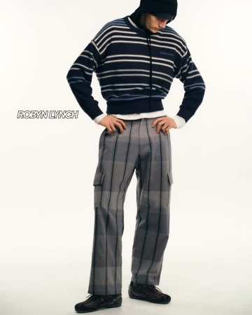 Male fashion model poses in a striped sweater and plaid pants in a full shot against a bright studio backdrop, showcasing contemporary menswear.
