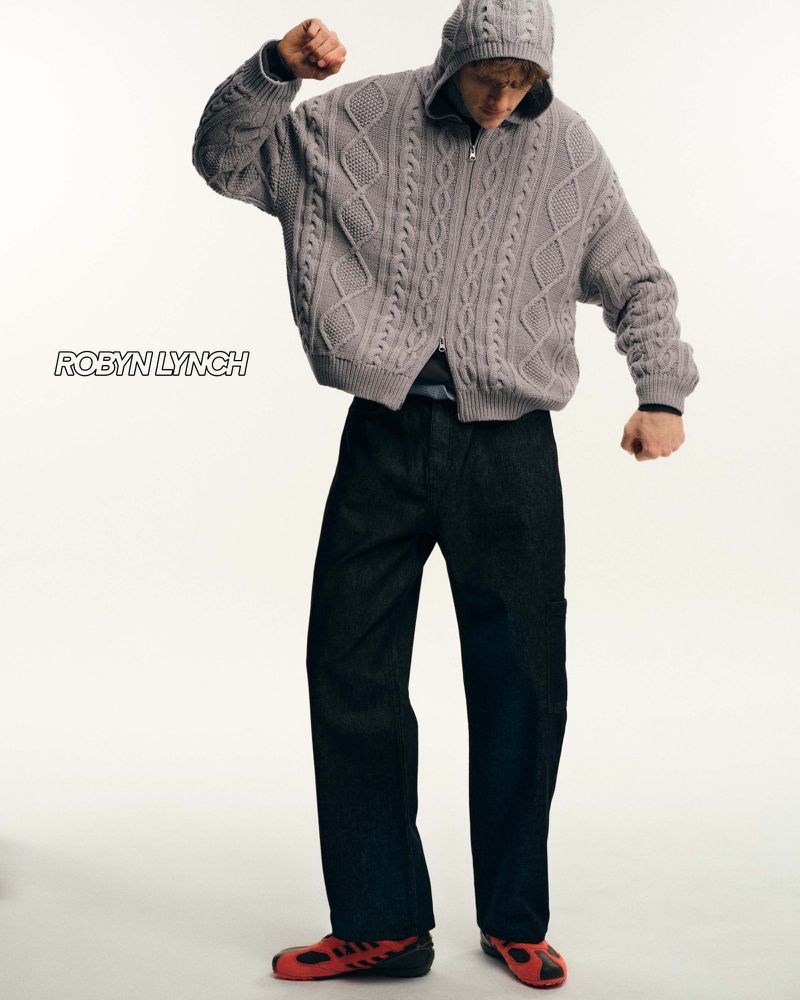 An adult male poses in a knitted, hooded zip-up sweater and black pants, with red shoes, standing in a photo studio against a neutral backdrop.
