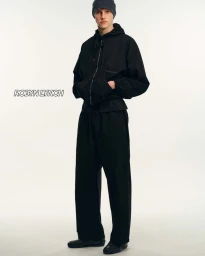 Model wearing a black zip-up hooded jacket and black trousers with a dark beanie against a plain white background