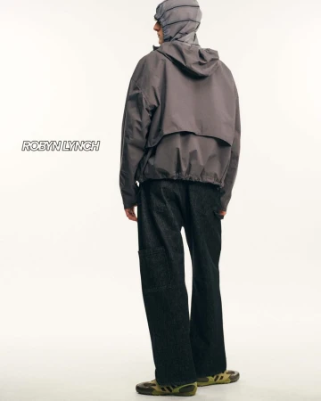 Model seen from behind wearing a grey hooded jacket and dark trousers against a plain white studio background.