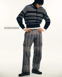 Model wearing a navy striped sweater, plaid trousers, and black beanie standing against a seamless white studio background