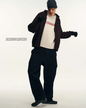 Model wearing a dark cable knit zip-up cardigan, cream t-shirt, dark beanie, and black trousers against a plain white background