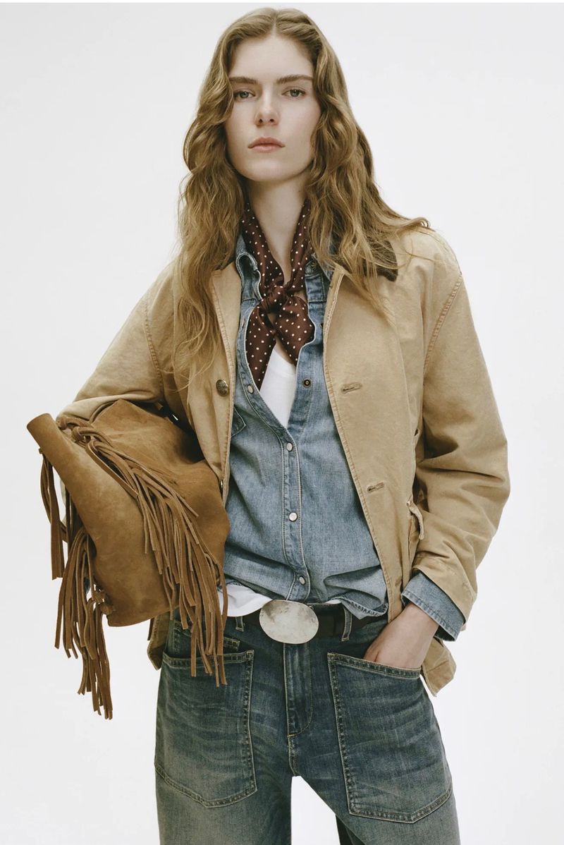 Woman wearing a denim shirt and jeans, with a tan jacket and suede fringe bag, posing in a studio with a neutral background.