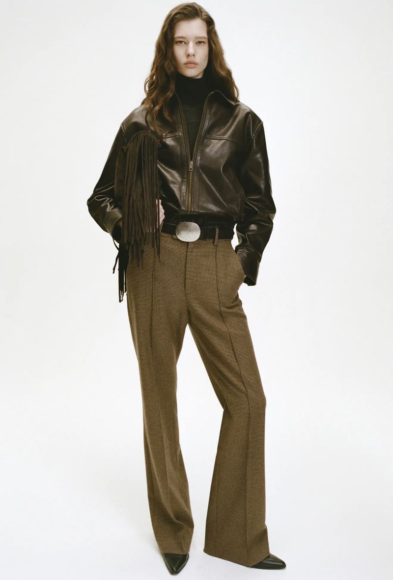 A full-length shot of a woman with wavy brown hair, wearing a black leather jacket with fringe, and wide-leg brown trousers.