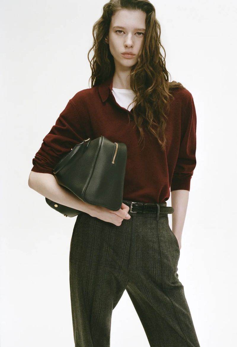 Portrait of a young woman in a burgundy sweater holding a black leather bag against a bright white background in a fashion editorial.