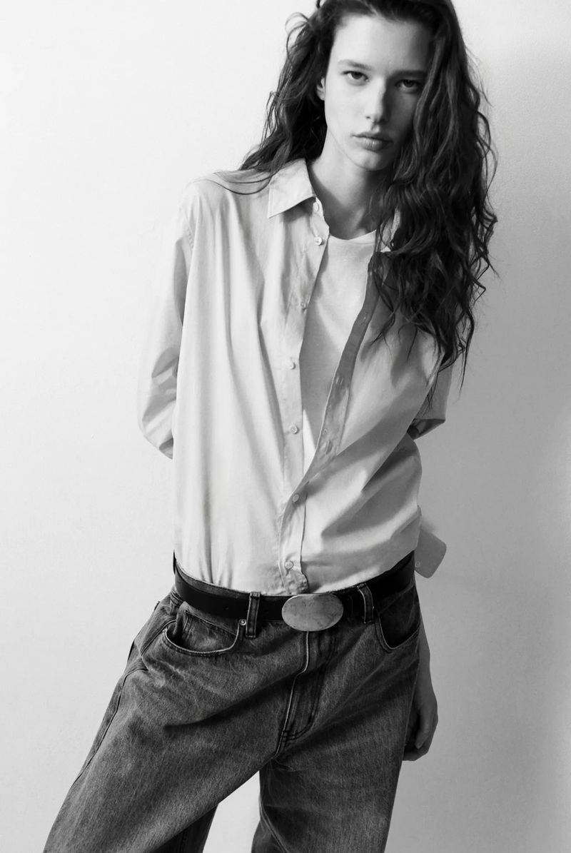 Monochrome portrait of a brunette woman in an open button-down shirt and jeans, set against a plain white backdrop in a studio setting.