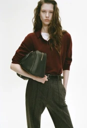 Model in burgundy polo shirt and dark trousers carrying a black leather handbag against a plain white background.