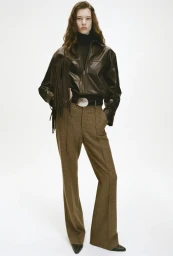 Model wearing a dark leather jacket and brown trousers against a white seamless background