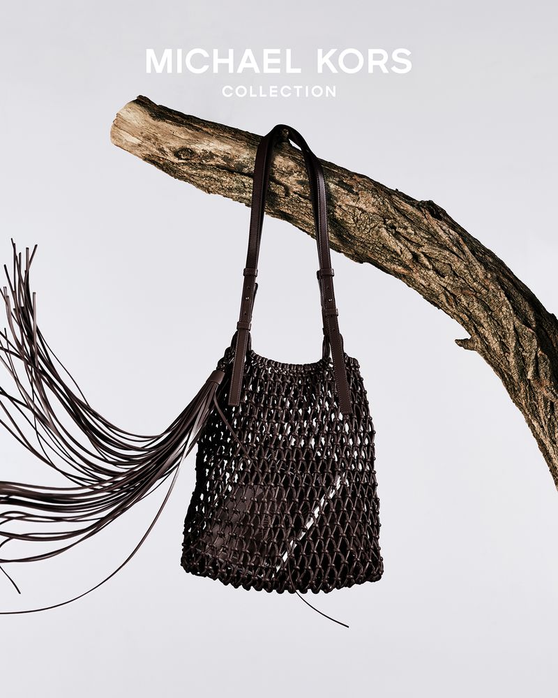 Michael Kors woven leather bag hanging from a tree branch against a soft white backdrop in a fashion product shot for luxury accessories.