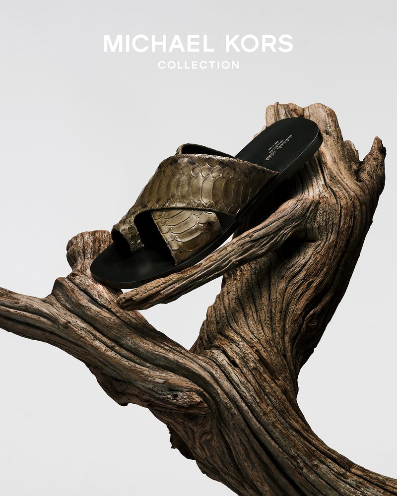 A sandal with a snakeskin pattern sits atop a textured tree branch against a clean white background, showcasing neutral tones and fashion.