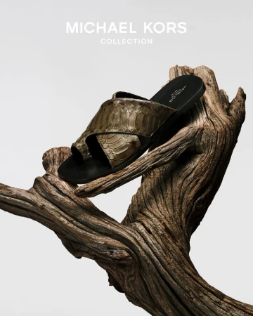 A sandal with a snakeskin pattern sits atop a textured tree branch against a clean white background, showcasing neutral tones and fashion.