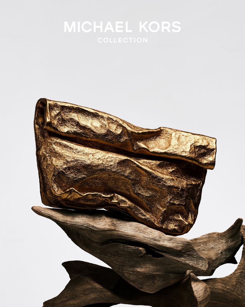 Gold Michael Kors clutch handbag rests atop driftwood fragments against a bright white background in a product studio shot.