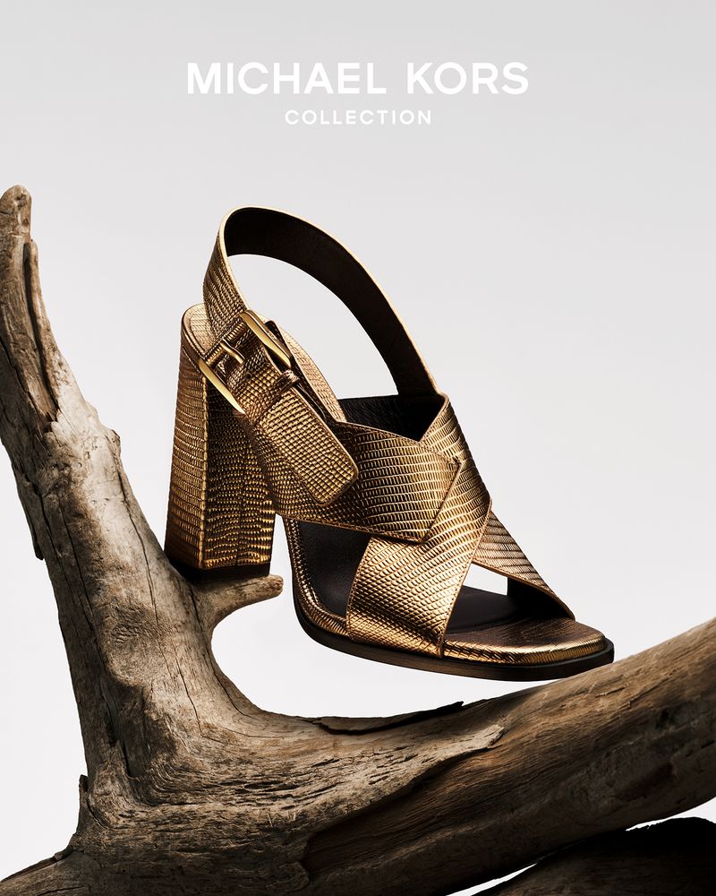 Gold-toned Michael Kors strappy sandal with a block heel sitting on a piece of driftwood against a neutral background.