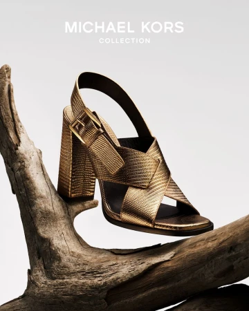 Gold-toned Michael Kors strappy sandal with a block heel sitting on a piece of driftwood against a neutral background.