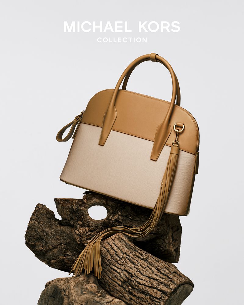 Michael Kors handbag in neutral tones displayed on a textured wood base, shot against a plain background in a studio setting.