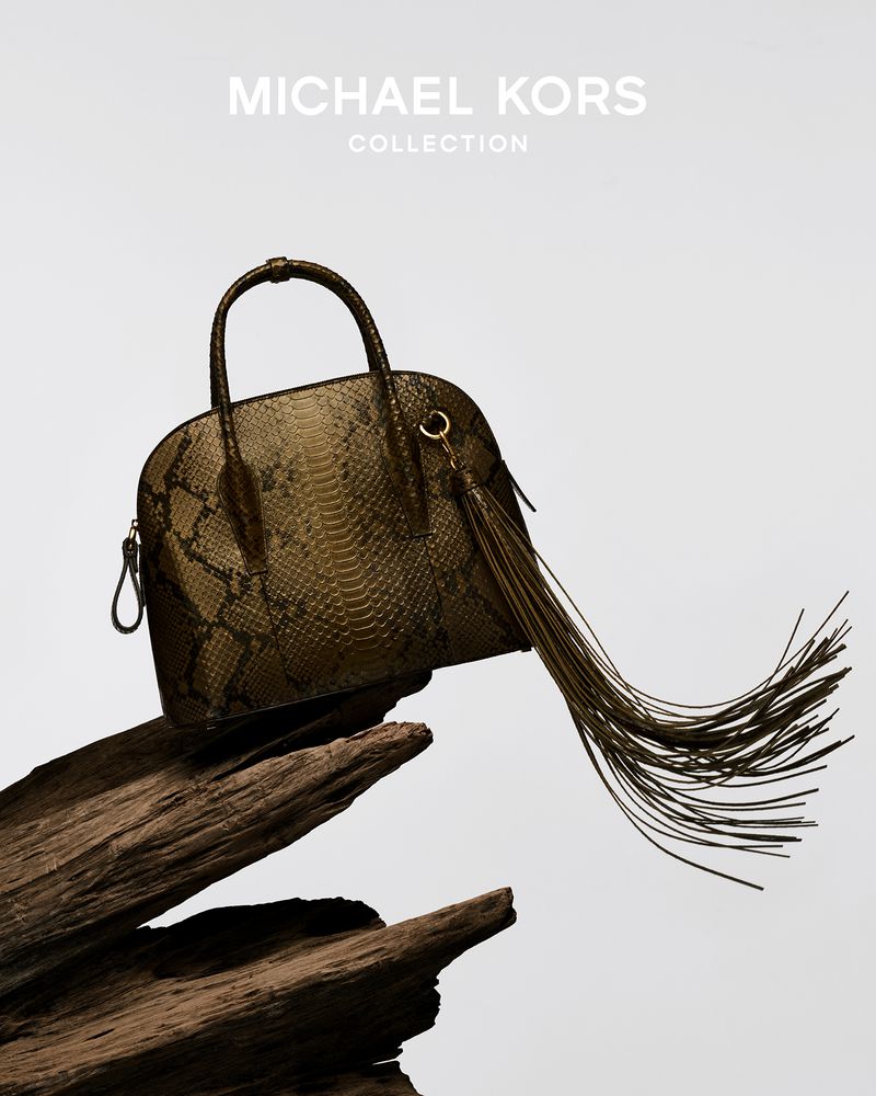 Olive snakeskin satchel handbag by Michael Kors, displayed on dark wooden surface against a light background in a studio setting.